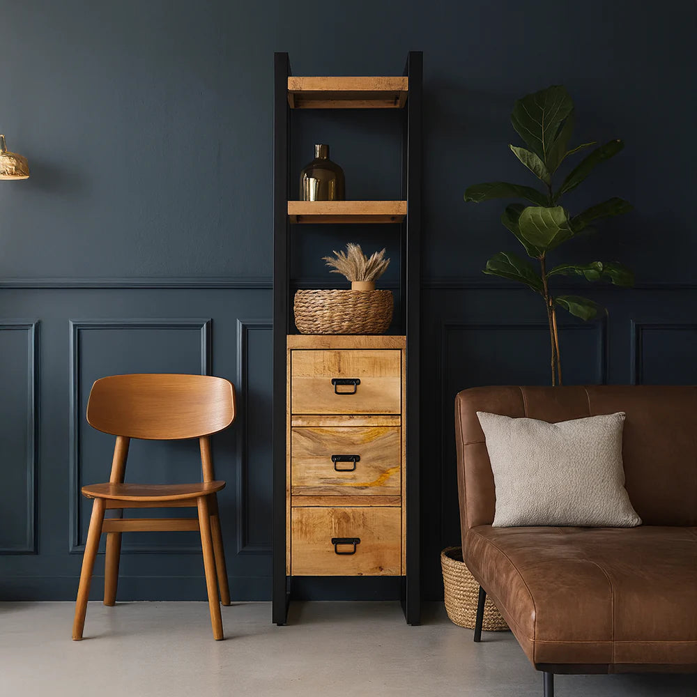 Sfeervolle presentatie van de mangohouten boekenkast Britt in een modern interieur met een donkerblauwe paneelwand, houten stoel en bruin lederen bank