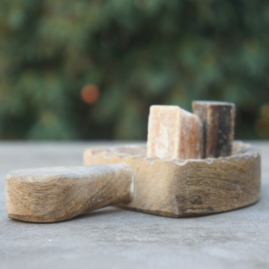 Rustiek schepvormig schaaltje van mangohout met een handvat, gepresenteerd met twee geurende amberblokjes tegen een sfeervolle, groene achtergrond van Rimisa.