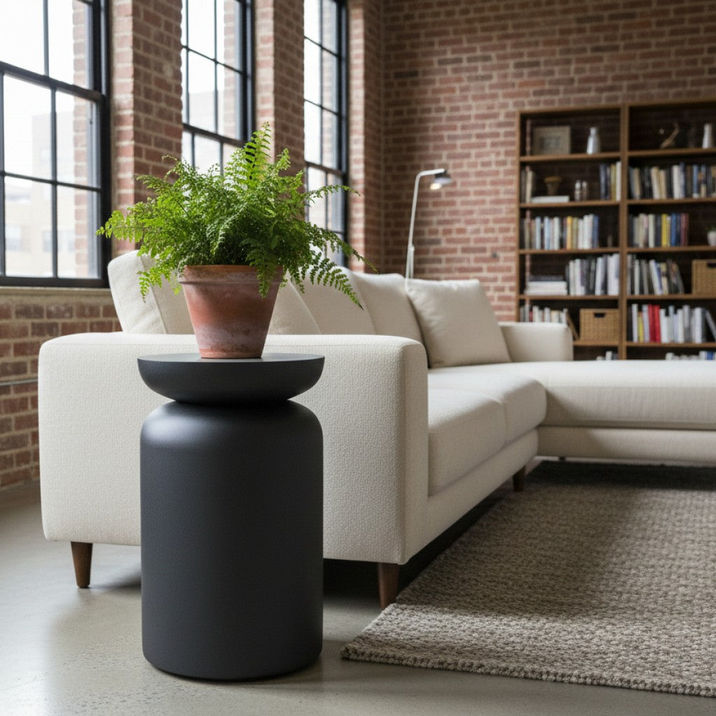 Zwarte ronde bijzettafel van By Moos met een groene varen plant naast een witte stoffen bank in een industrieel interieur.